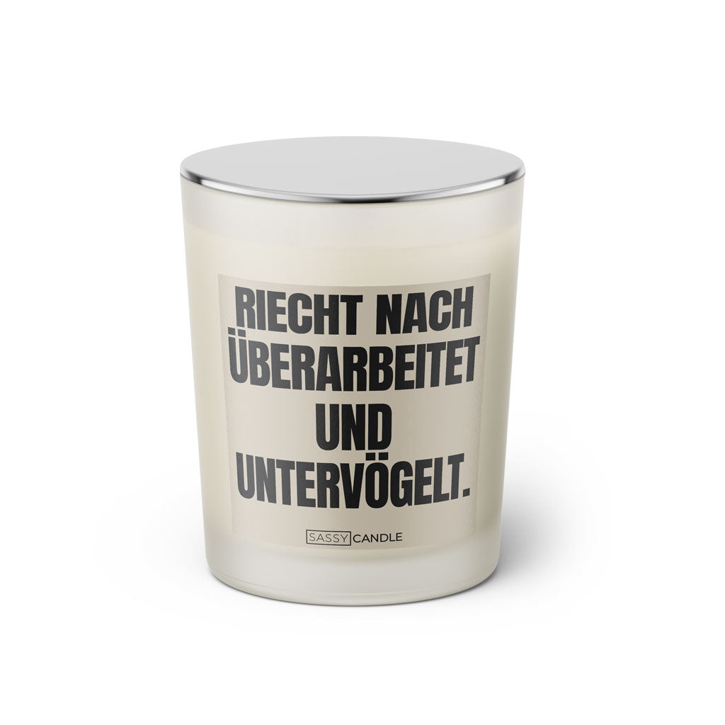 Duftkerze mit witzigem Spruch: Riecht nach untervögelt und überarbeitet. Farbe beige und schwarze Schrift. Kerzenglas mit silbernem Deckel von SassyCandle.