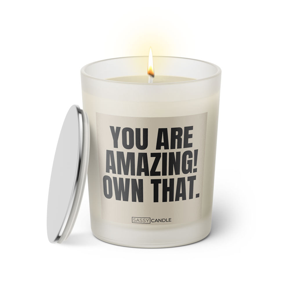 Duftkerze mit witzigem Spruch: You are amazing! Own that. Farbe beige und schwarze Schrift. Kerzenglas mit silbernem Deckel von SassyCandle.