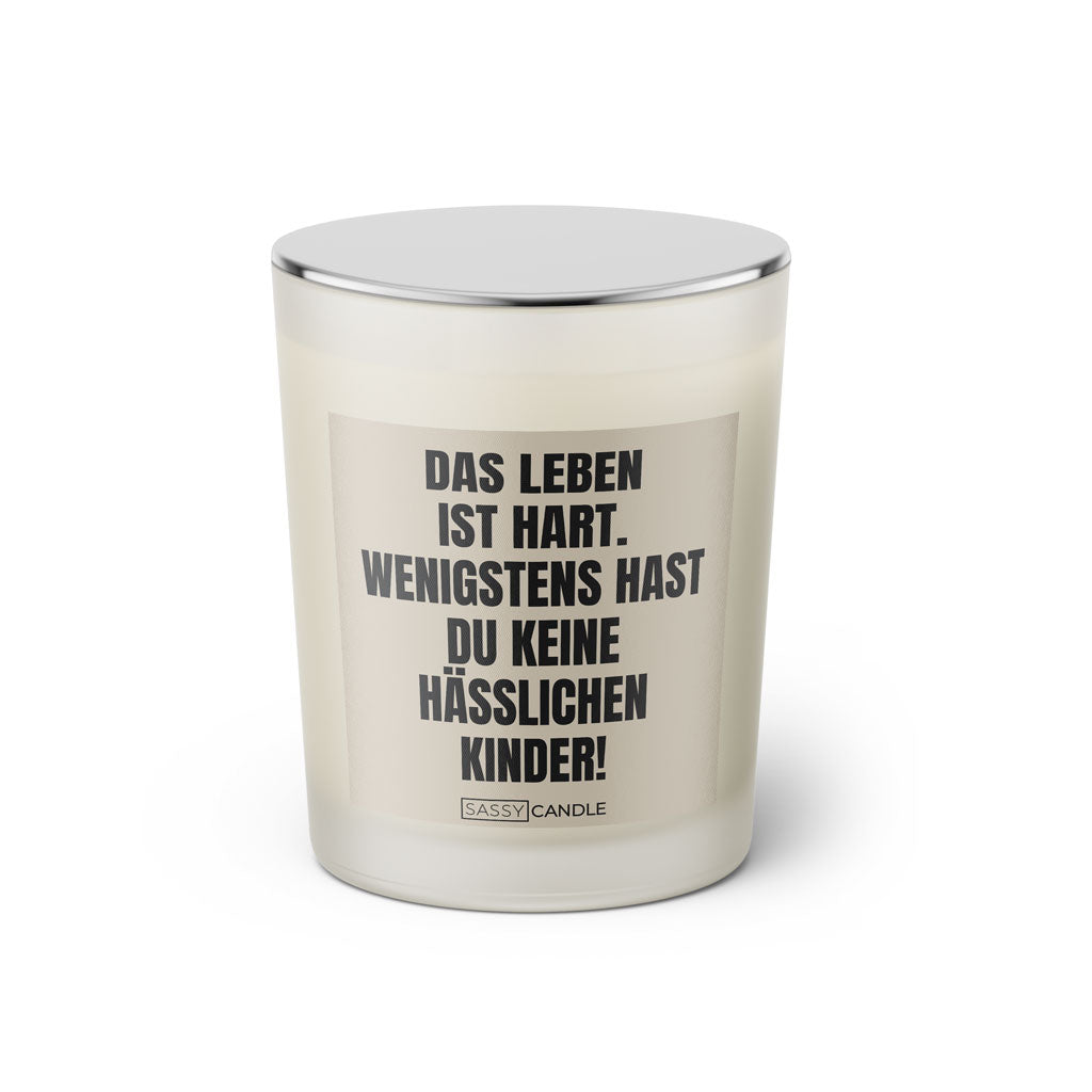 Duftkerze mit witzigem Spruch: Das Leben ist hart. Wenigstens hast du keine hässlichen Kinder. Farbe beige und schwarze Schrift. Kerzenglas mit silbernem Deckel von SassyCandle